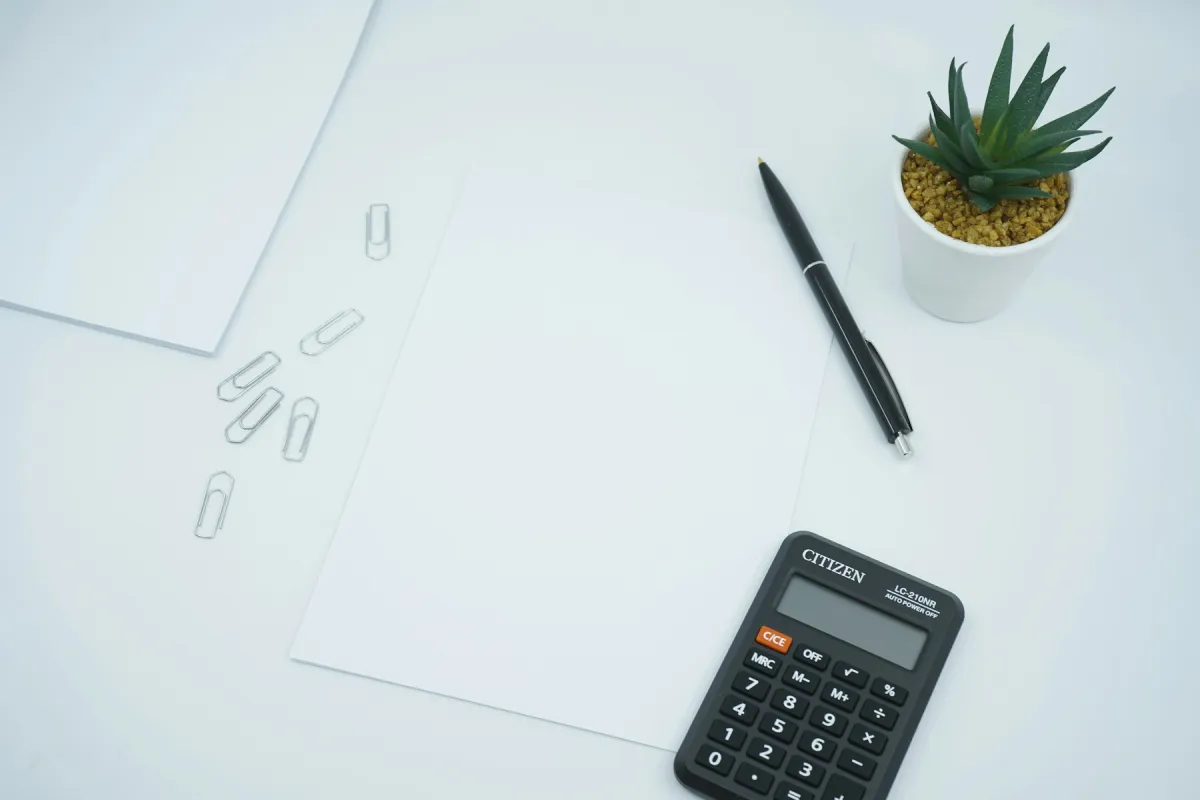 A calculator, black pen, paper clips and blank paper on a white desk beside a potted plant.