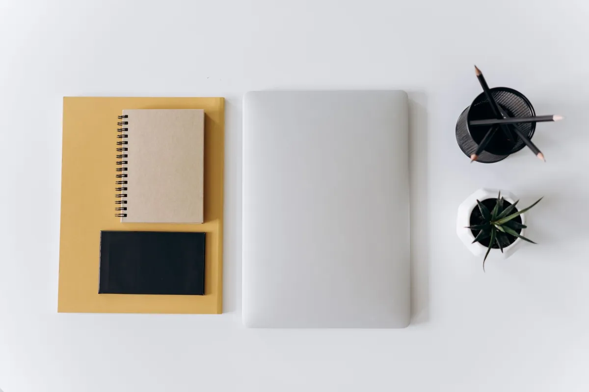 Closed silver laptop beside notebooks, pencils and a small plant on a white desk.