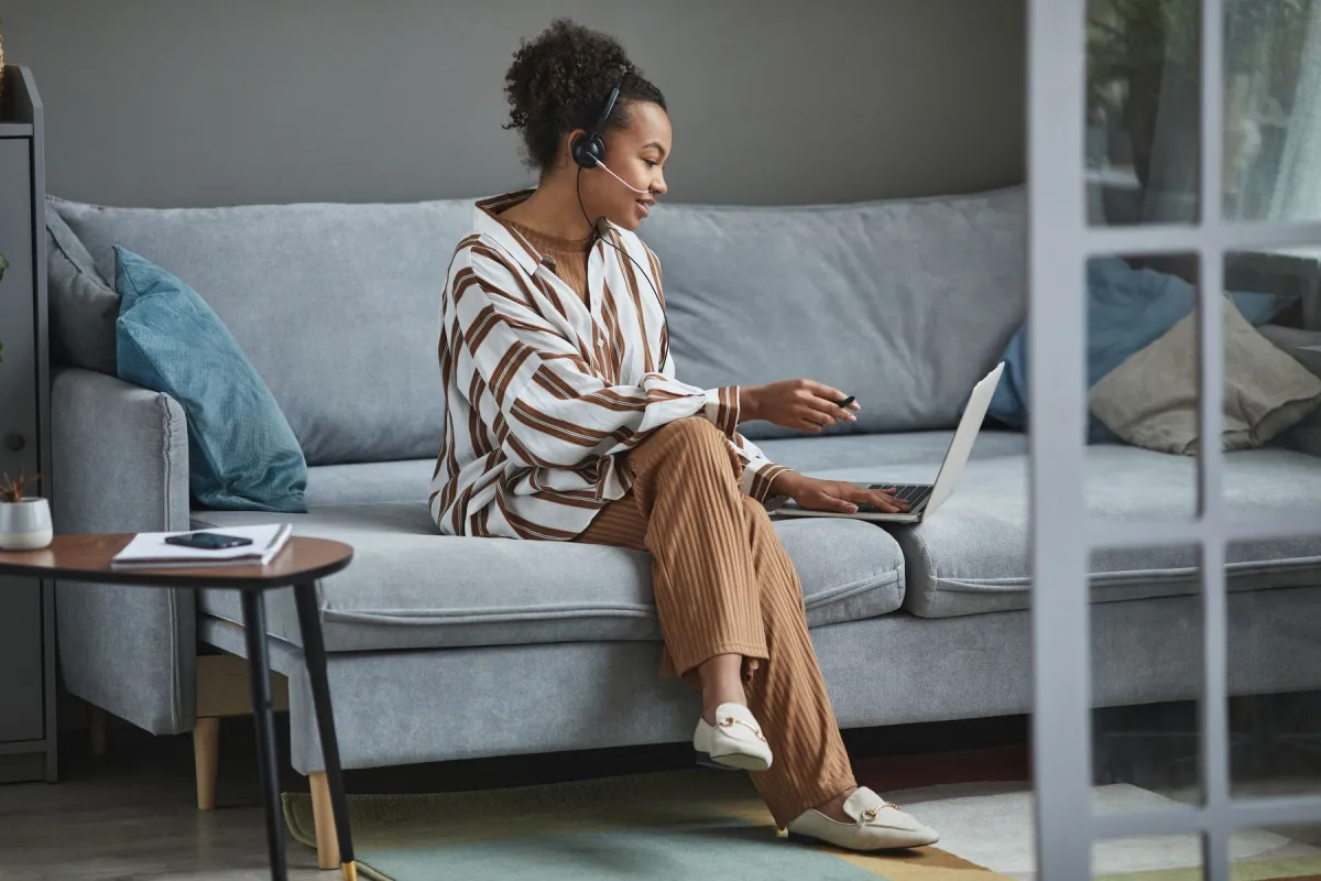 A woman wearing a boom-microphone headset uses a laptop while seated on a sofa.
