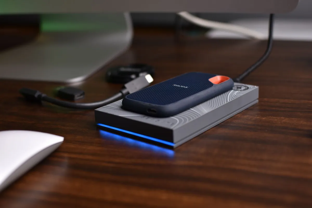 A portable SanDisk SSD with an unplugged front socket rests on a silver enclosure on a wooden desk.