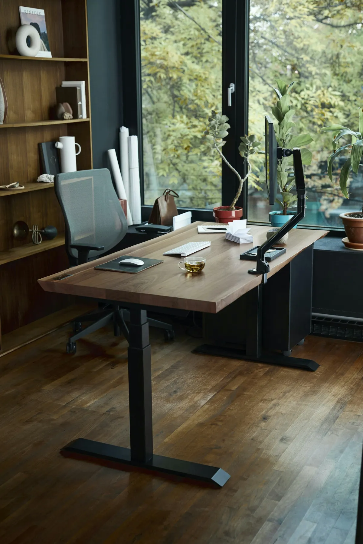 Wooden desk with a monitor on an arm, white keyboard and mouse beside a leafy window.