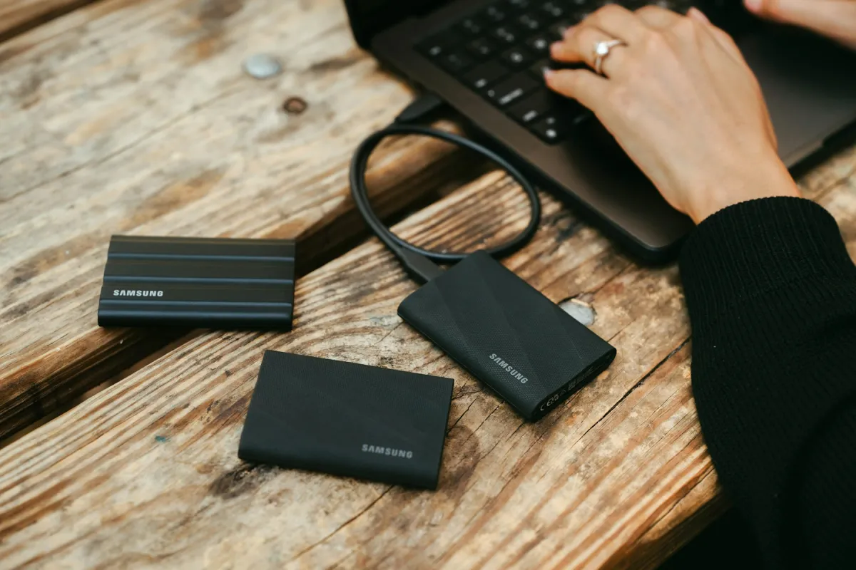 Person using a laptop beside three black portable storage drives on a wooden table.