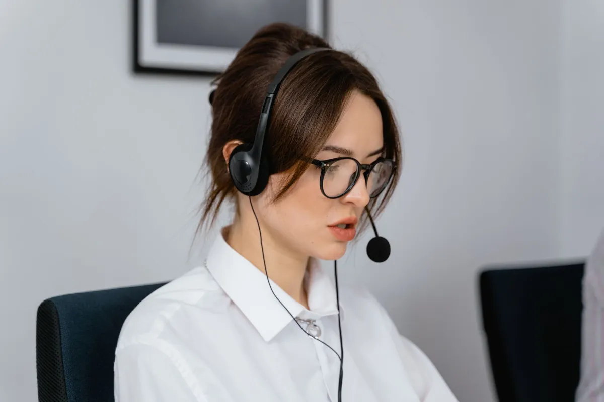 A woman wearing glasses and a wired headset with a foam-covered microphone boom sits at a computer.