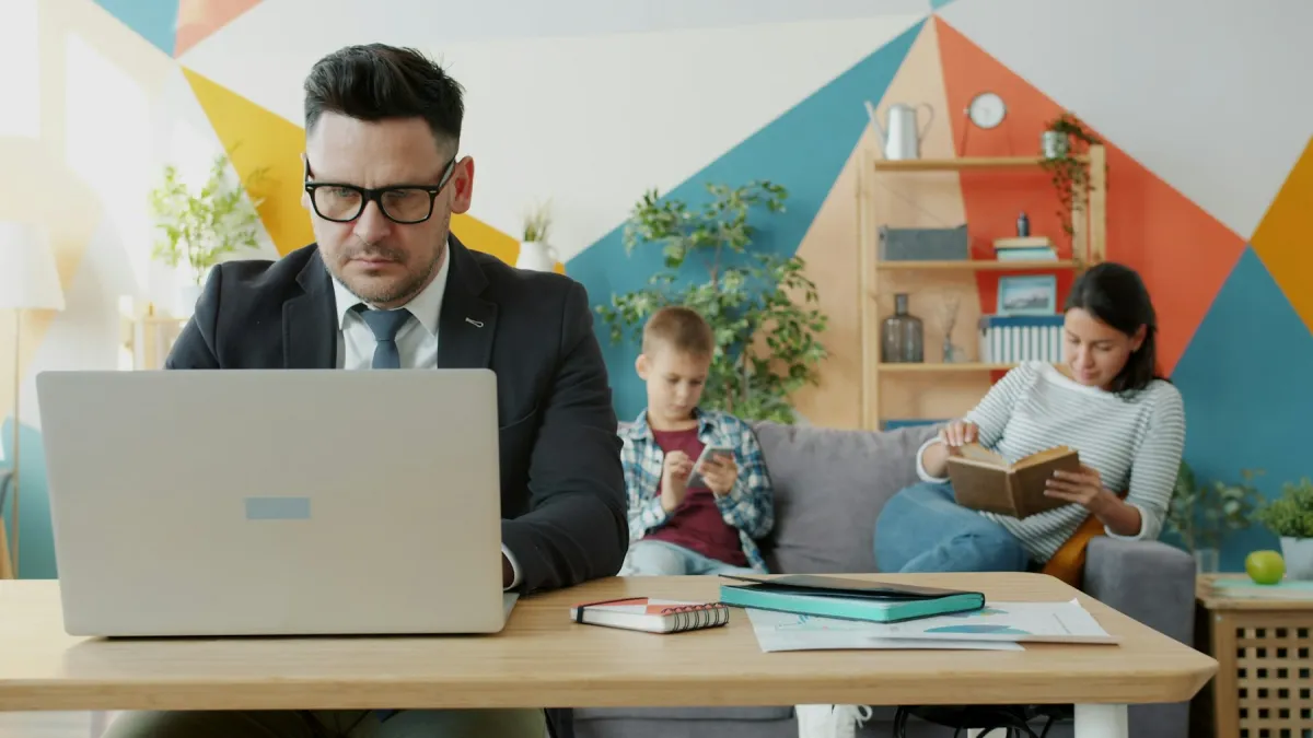 Man working on a laptop at a table while a child uses a phone and a woman reads on the sofa behind him.