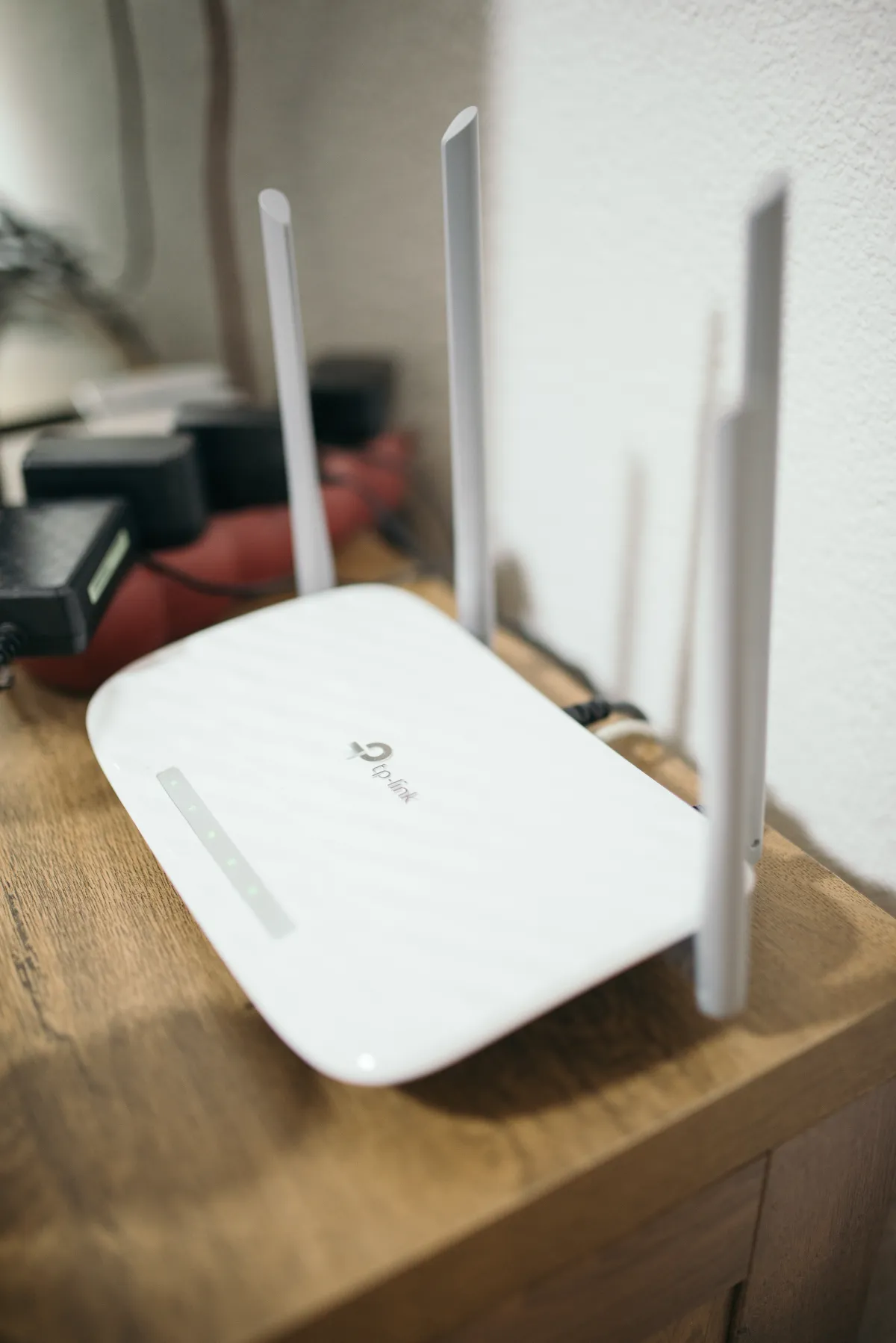 White TP-Link router with upright antennas and illuminated indicators on a wooden table beside a wall.