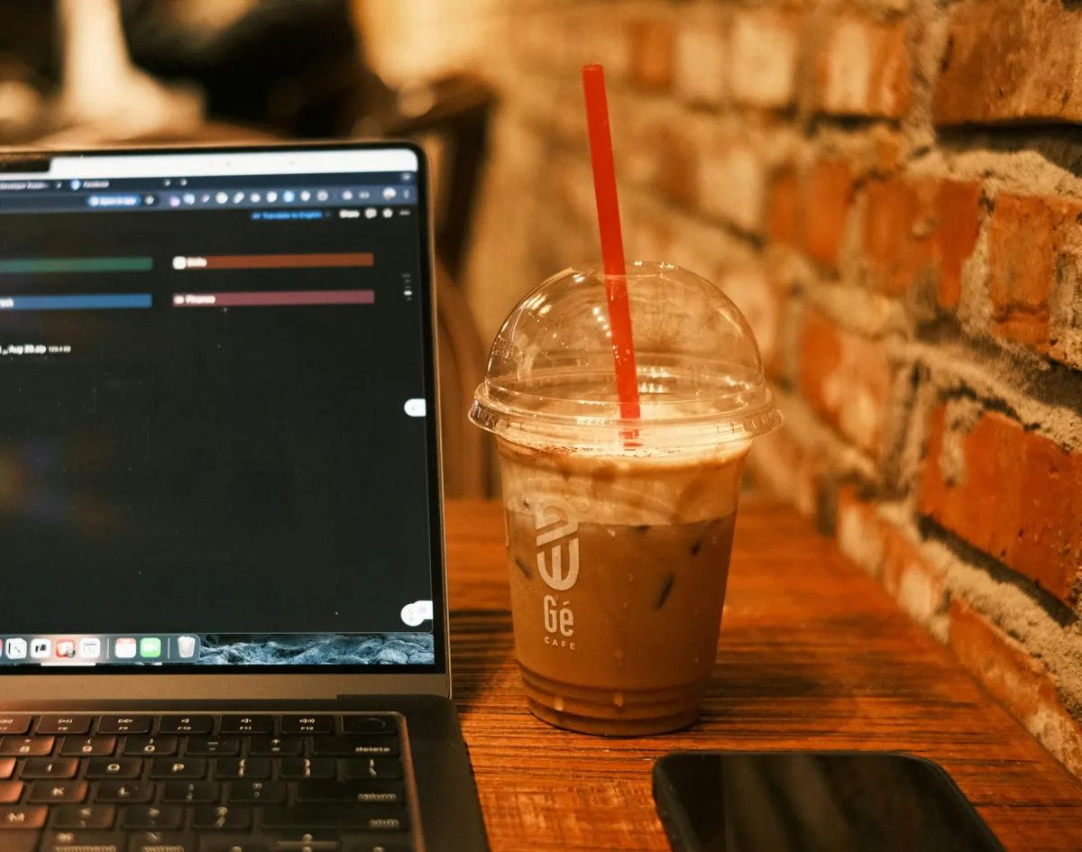 Laptop beside an iced coffee on a wooden café table against a brick wall.