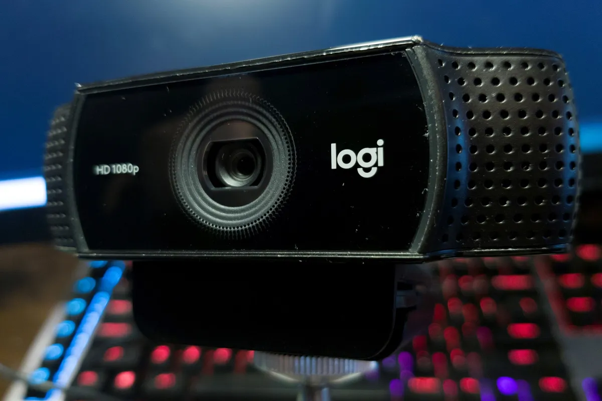 Close-up of a black Logitech webcam with its lens facing forward, above a blurred illuminated keyboard.