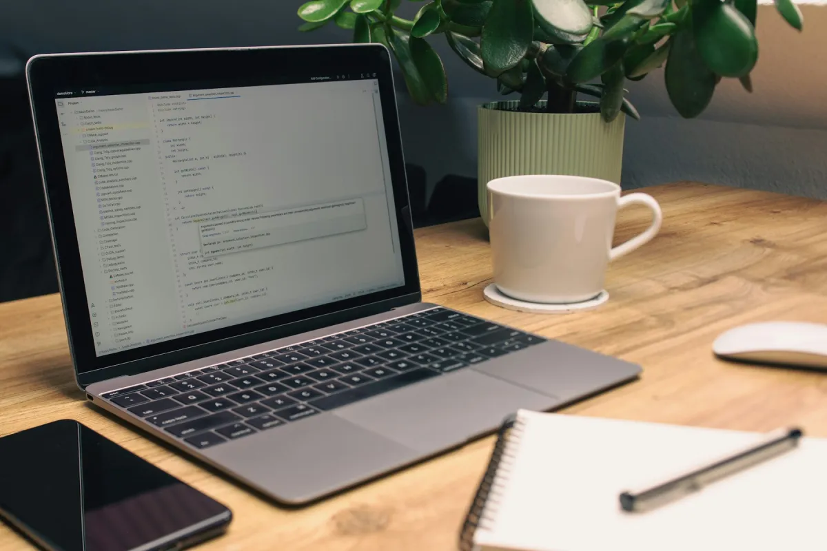 Laptop showing a code editor on a wooden desk beside a notebook, phone, white mug and potted plant.