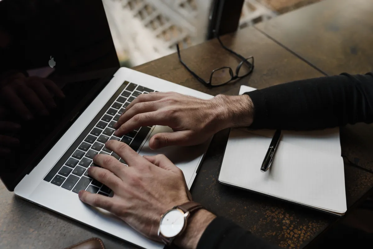 Hands on a laptop keyboard beside a lined notebook, pen and glasses on a wooden desk.