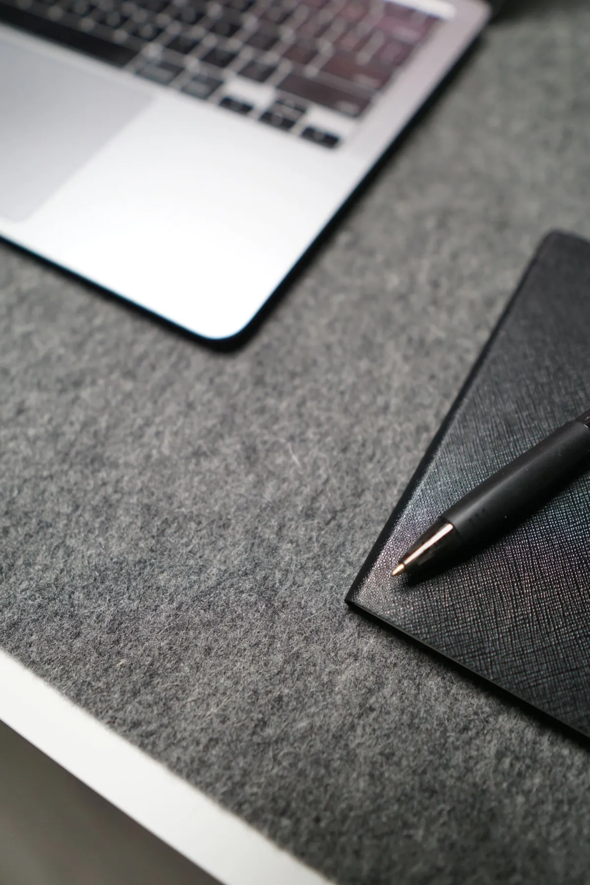 A silver laptop beside a closed dark notebook and pen on a gray desk surface.