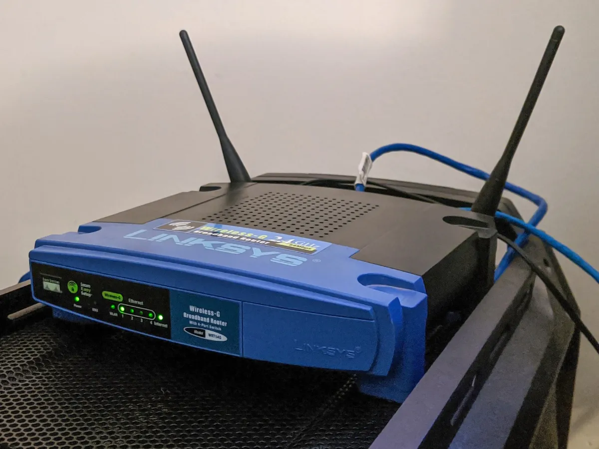 Blue and black Linksys WRT54G router with two antennas, front indicator lights and a blue cable behind it.