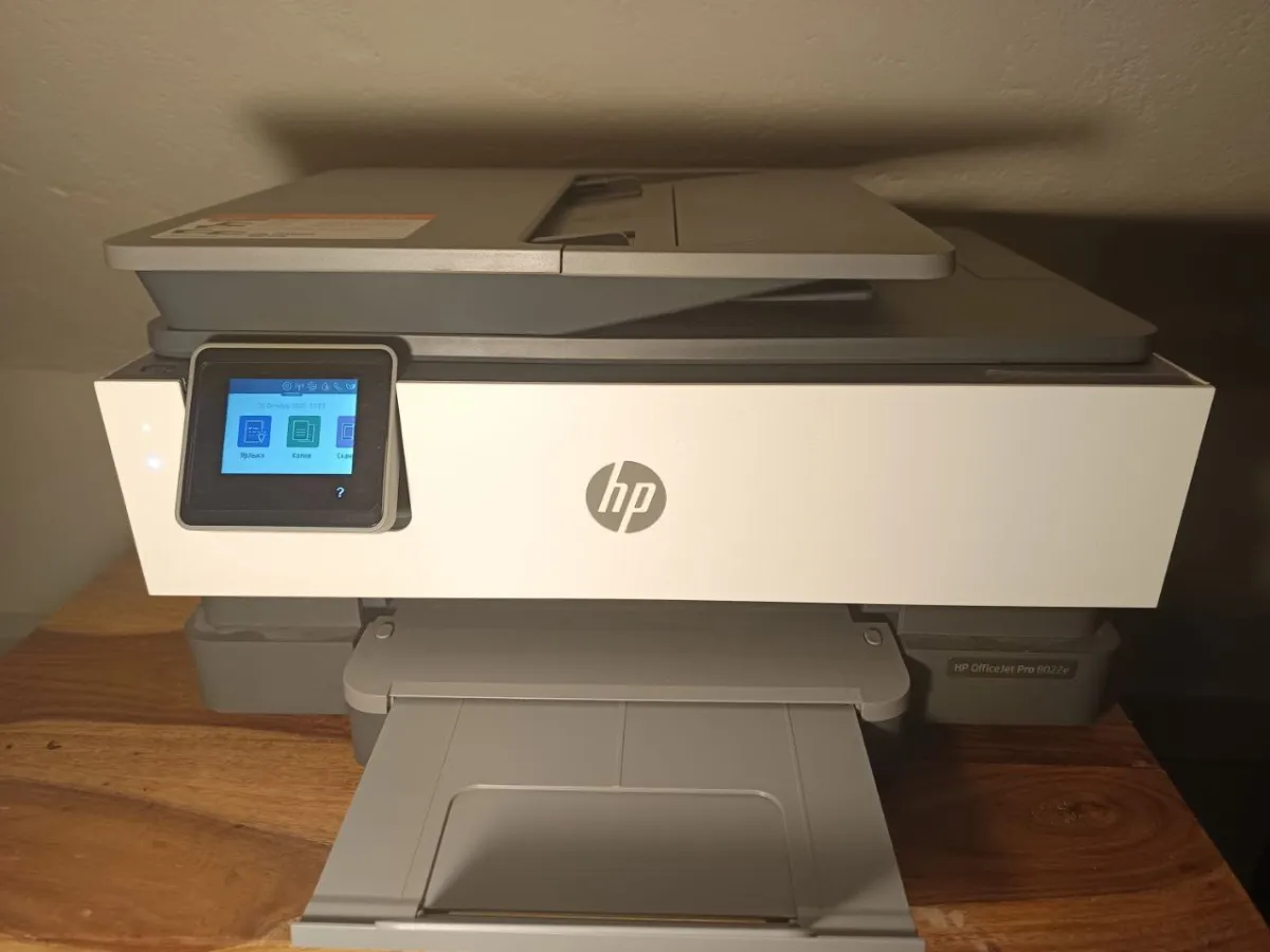 White HP OfficeJet Pro 8022e printer on a wooden surface with its touchscreen lit and paper output tray extended.