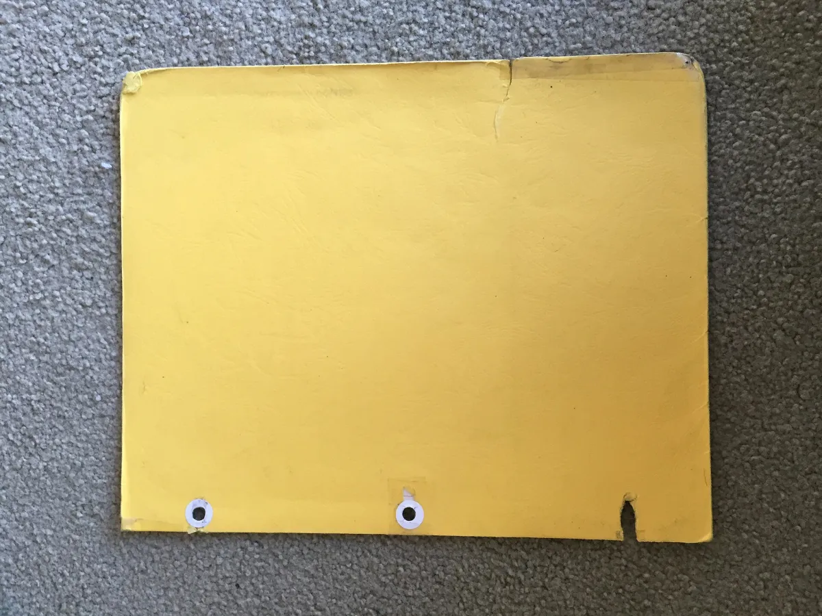 Worn yellow paper file folder with reinforced holes lying on grey carpet.