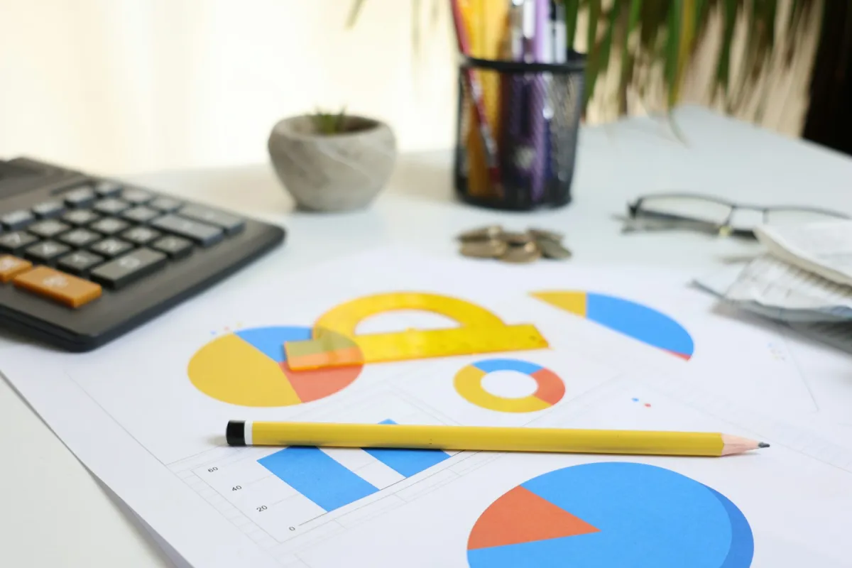 Printed charts with a yellow pencil and protractor on a desk beside a calculator, coins and glasses.