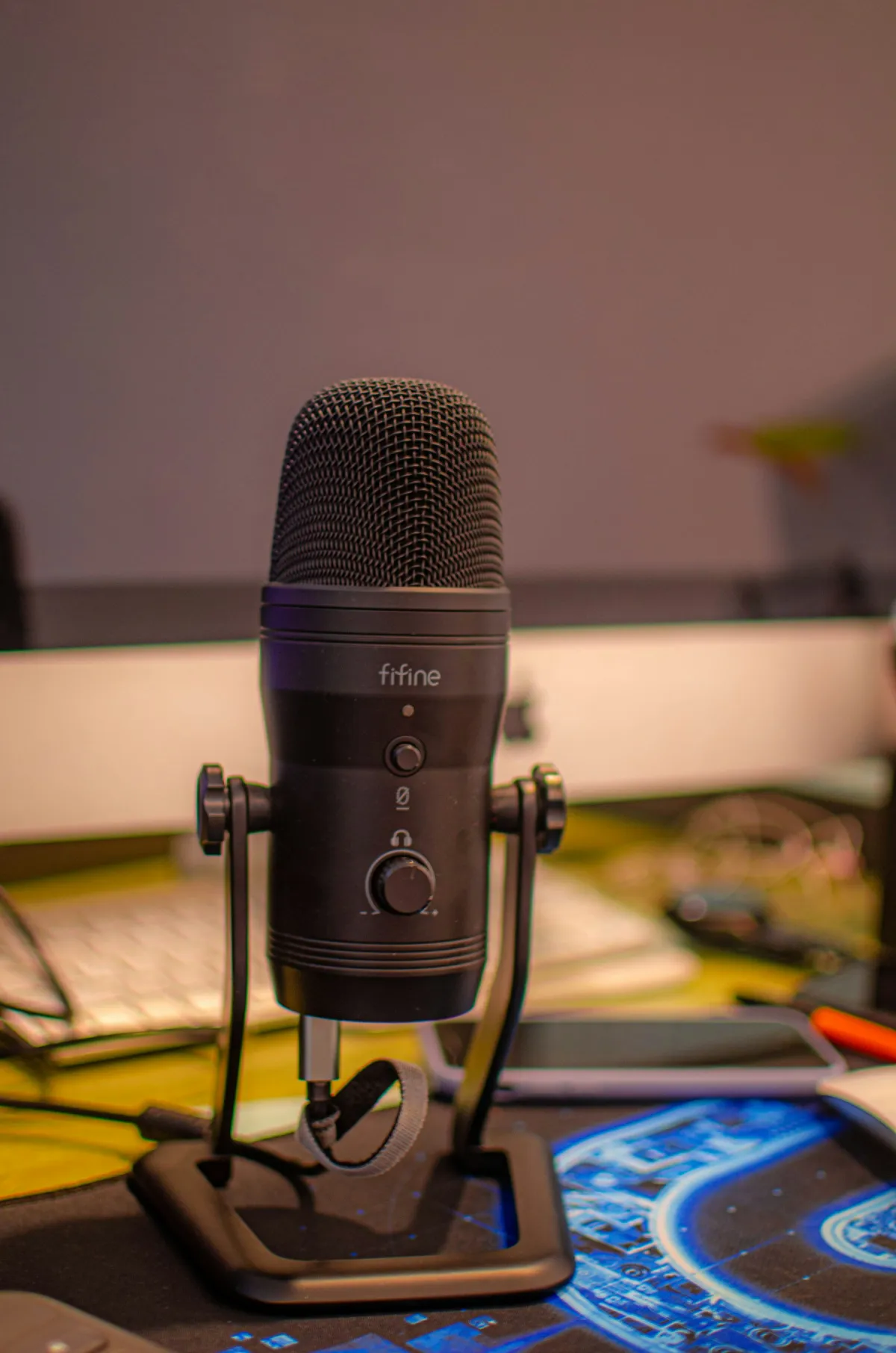 Black desktop microphone on a stand with a keyboard, phone and desk mat behind it.
