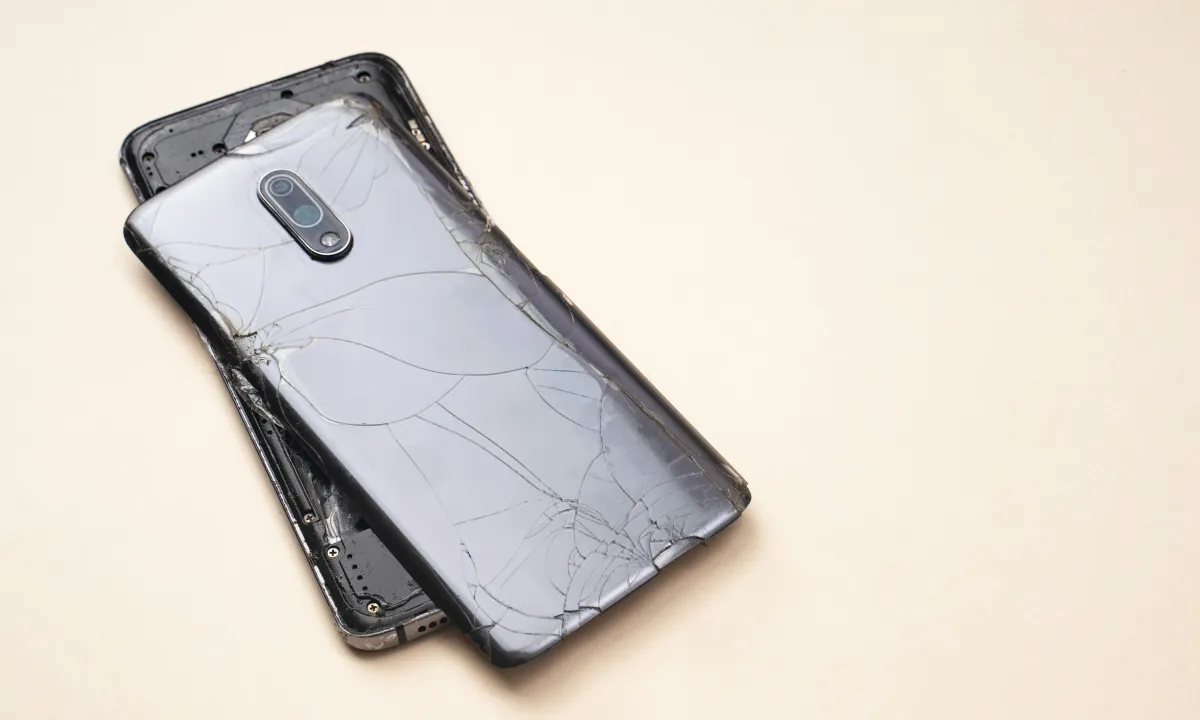 A damaged smartphone with cracked rear glass and its back partly separated, lying on a pale surface.