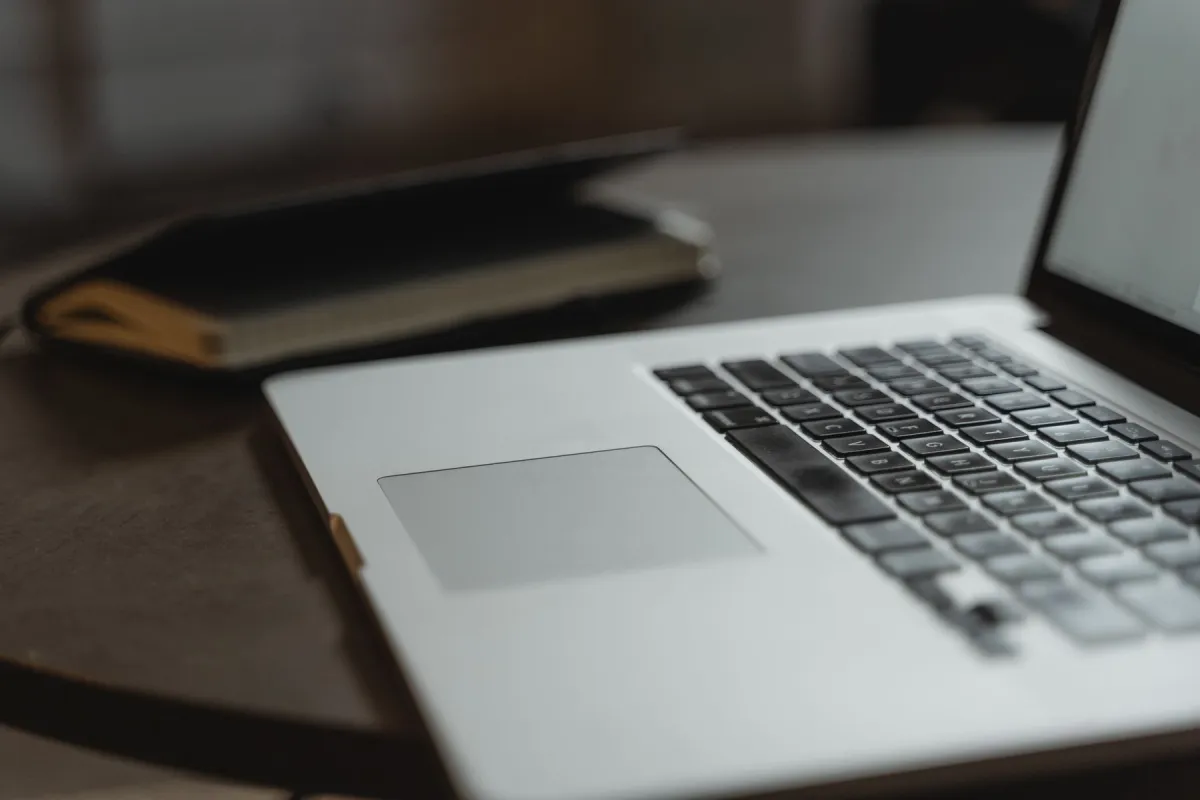Close view of a silver laptop keyboard and trackpad on a wooden desk, with a notebook behind it.