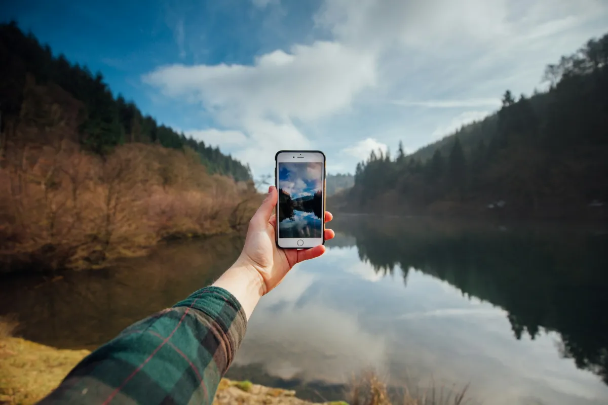A hand holds an iPhone framing a wooded lake beneath a blue sky.