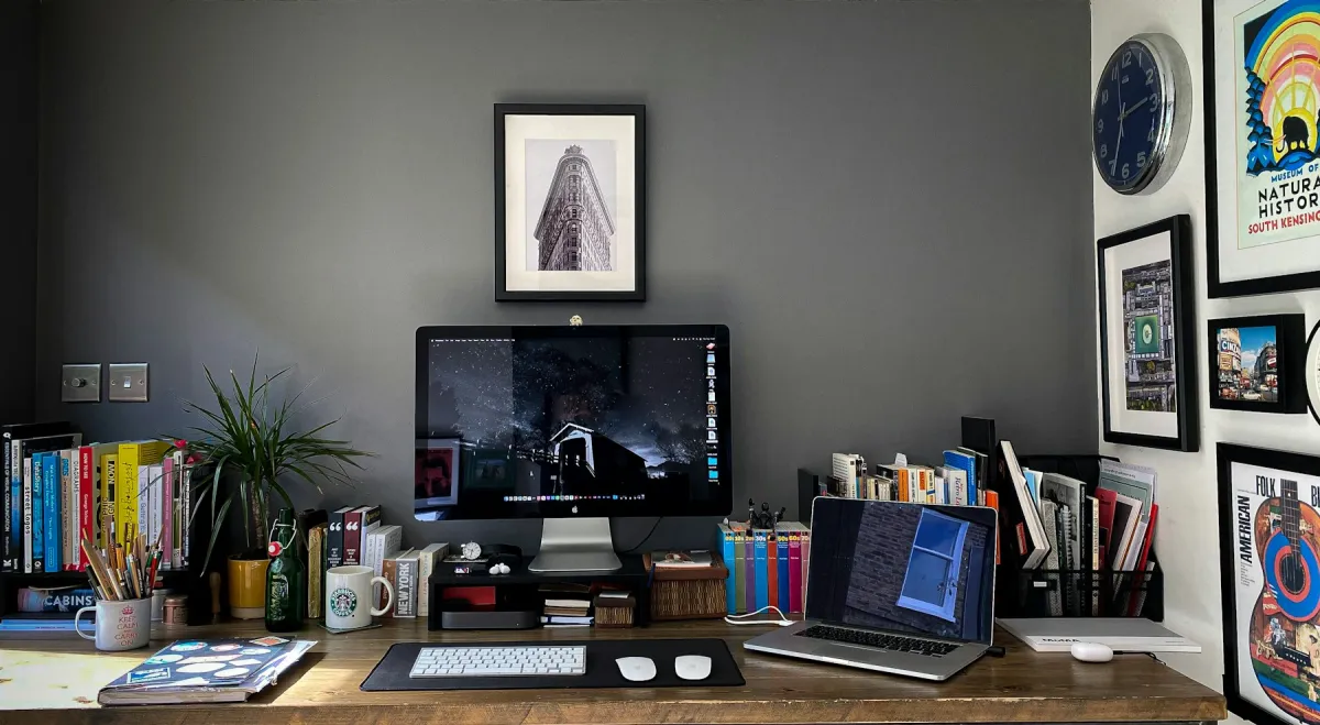 Desktop computer and open laptop on a wooden home-office desk surrounded by books and framed artwork.