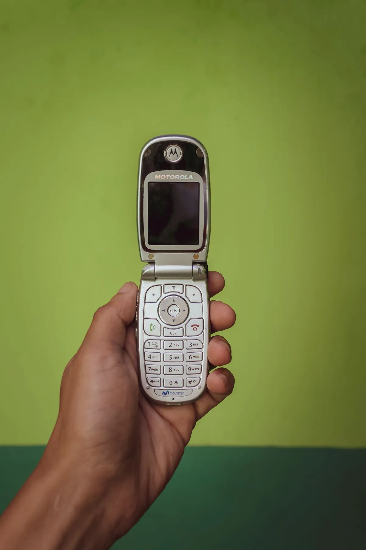 A hand holding an older Motorola flip phone against a green background.