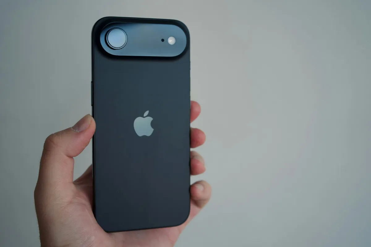 A hand holds the back of a dark iPhone against a pale background.