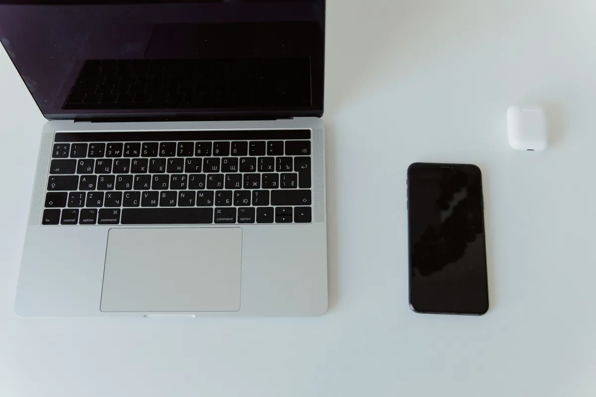 A MacBook, dark-screen iPhone and white wireless earbud case on a white tabletop.