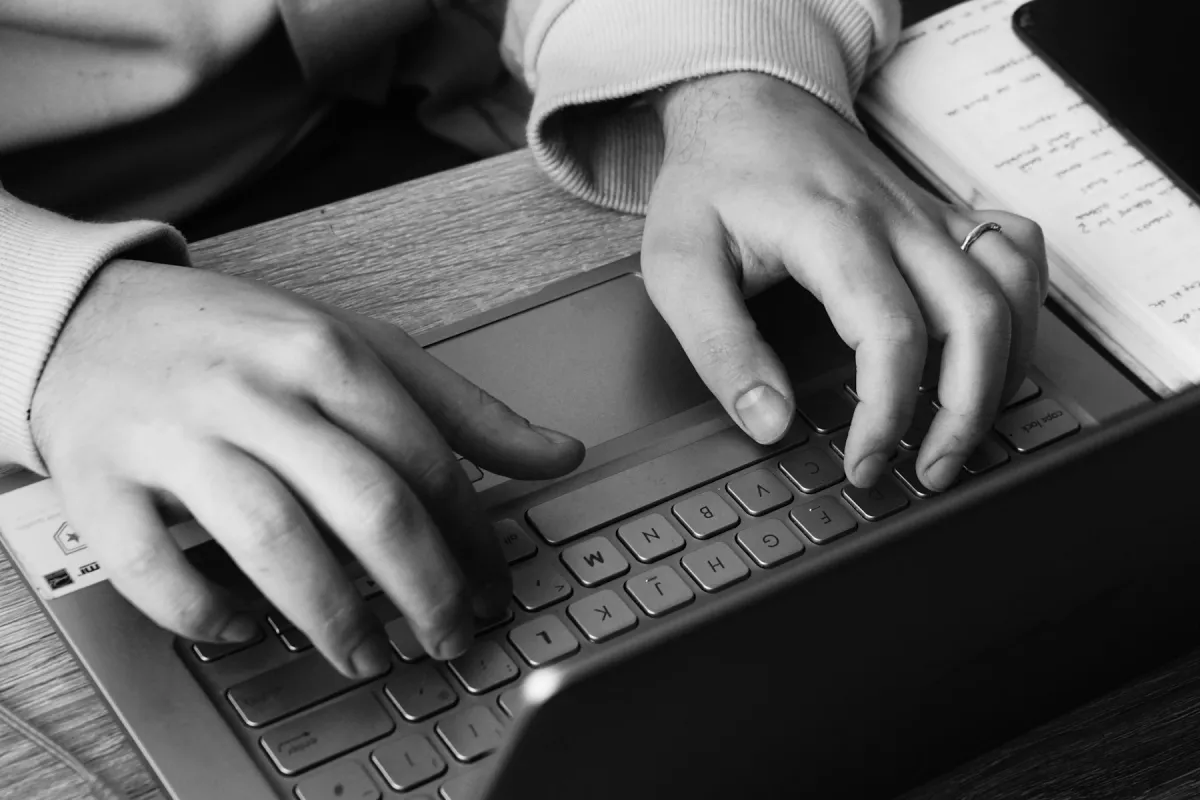 Black-and-white photograph of hands typing on a laptop keyboard beside an open notebook.
