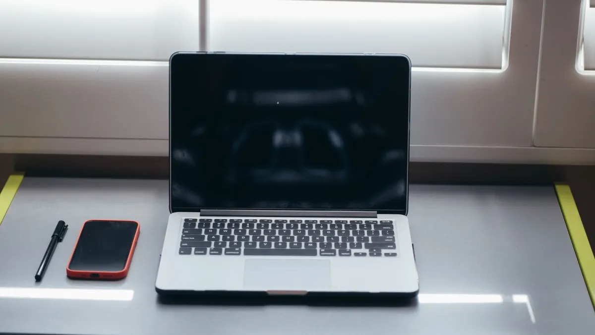 A laptop with a dark screen, a red-cased smartphone and a black pen on a desk beside a window.