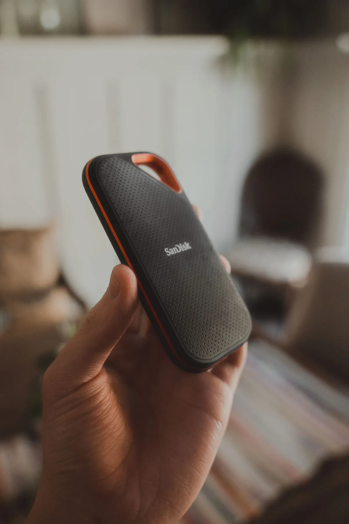 A hand holding a black and orange SanDisk portable SSD indoors.