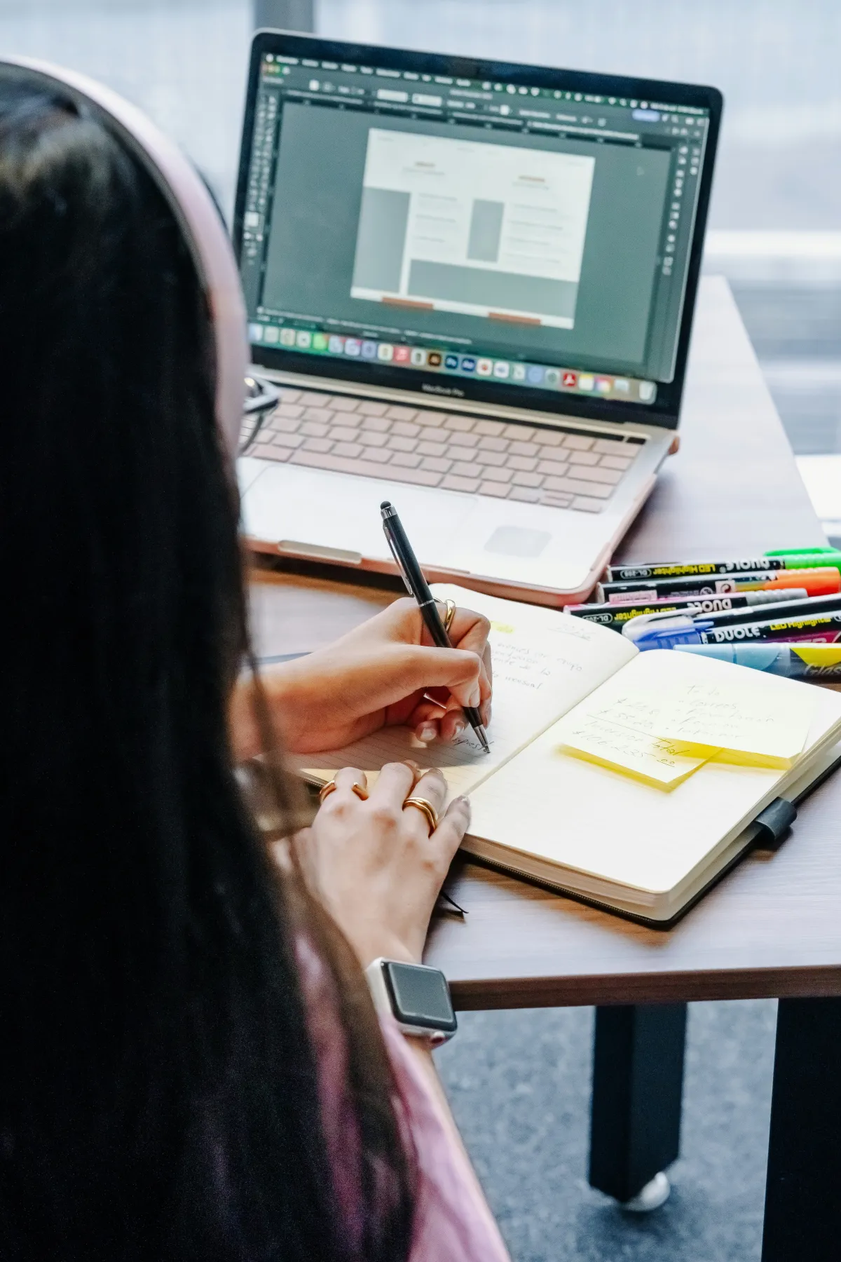 A person writes notes beside a laptop and a row of colorful pens on a desk.