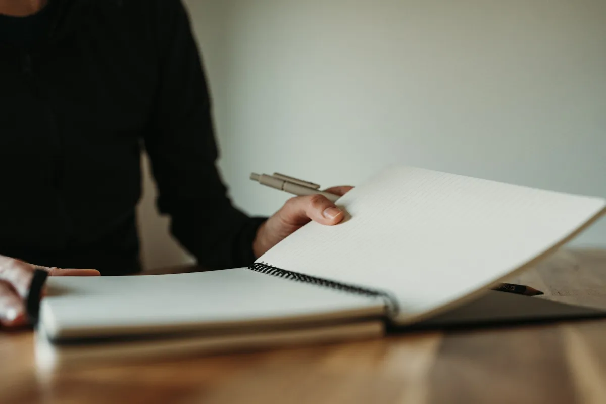 A person holding a pen and turning a lined page in a spiral notebook on a wooden table.