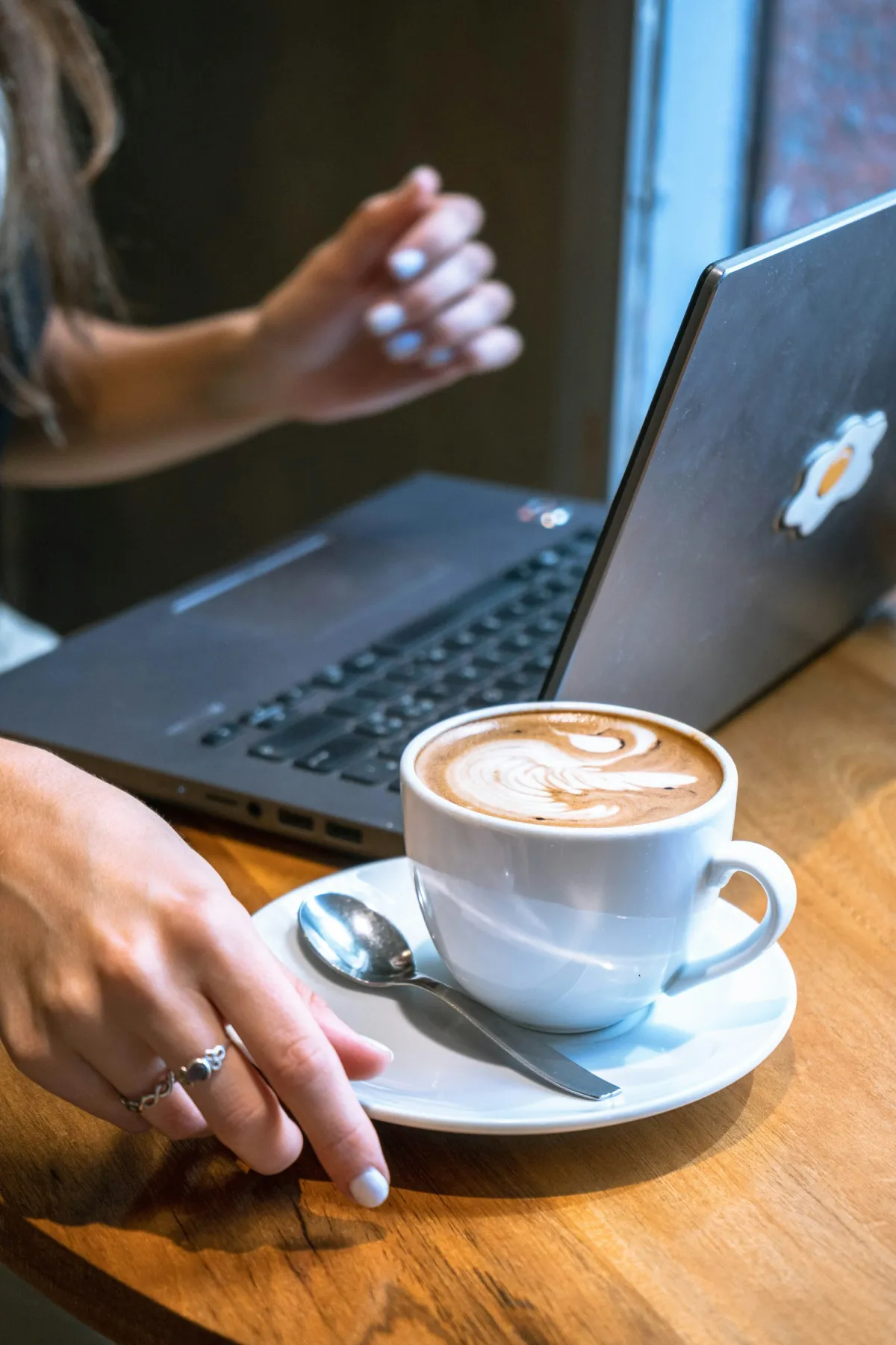 Coffee with latte art beside an open laptop and a person’s hands at a wooden cafe table.
