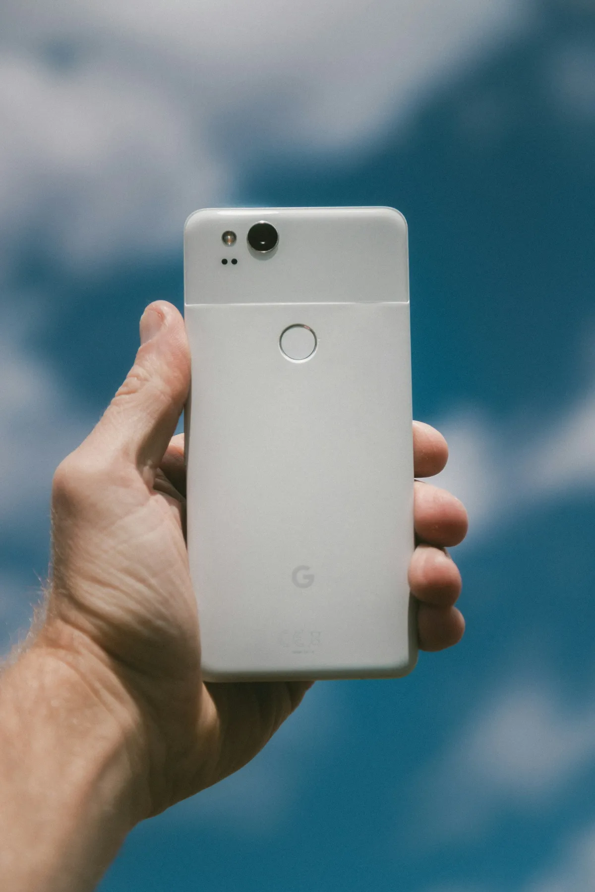 A hand holding the white back of an older Google Pixel phone against a blurred blue background.