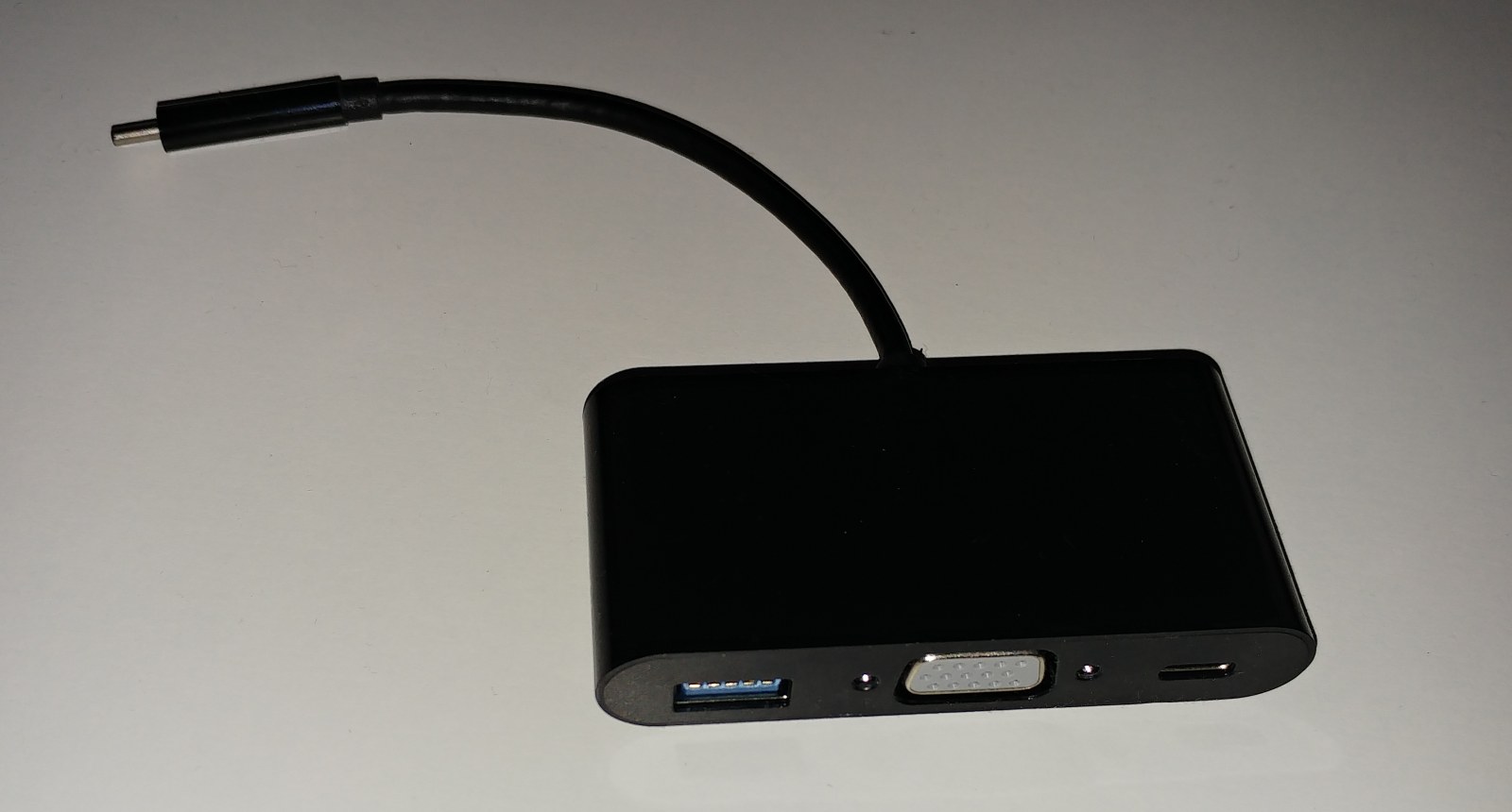 Black USB-C hub with captive host plug, USB-A socket, VGA socket and USB-C socket; no readable port-function labels appear beside the sockets.